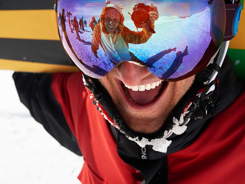 Person taking a photo reflected in the goggles of a snowboarder