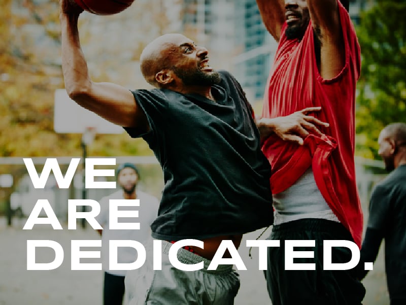 A man taking a shot in basketball while another man stands in his way. The words on the image read: We are dedicated.