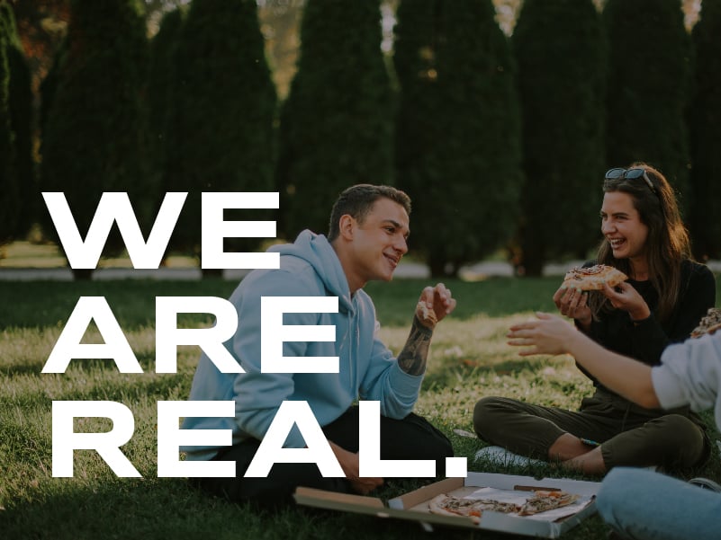 Three people sitting on the grass outside laughing and eating pizza. The words on the image read: We are real.