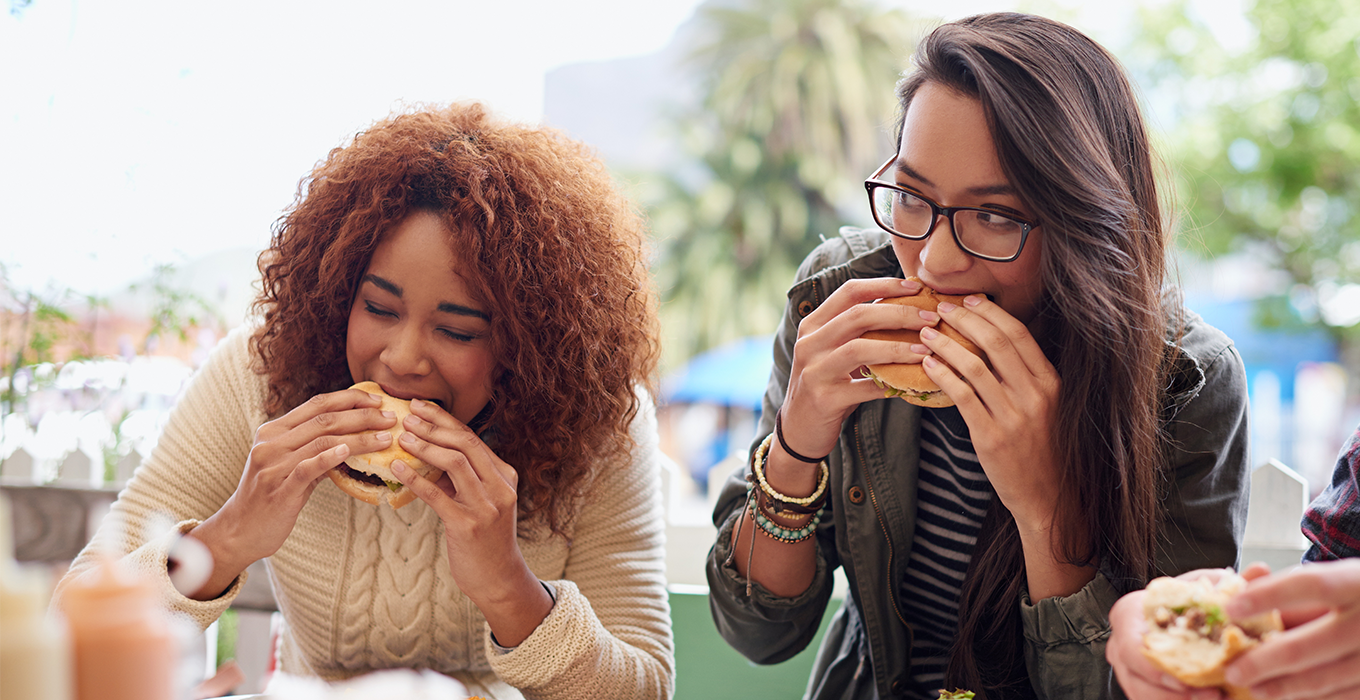 Two women are seated outside, eating hamburgers at a restaurant. 