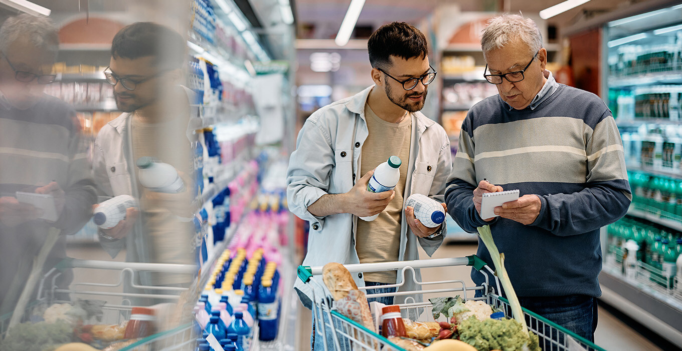 Two adult men compare two bottled beverages against their list while grocery shopping. 