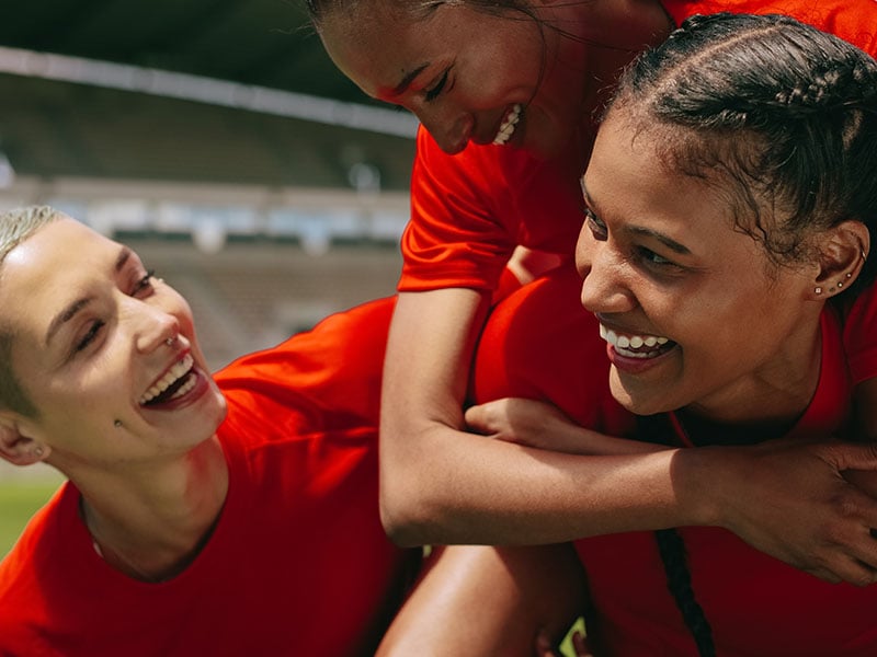 Three female teammates celebrating on a soccer pitch
