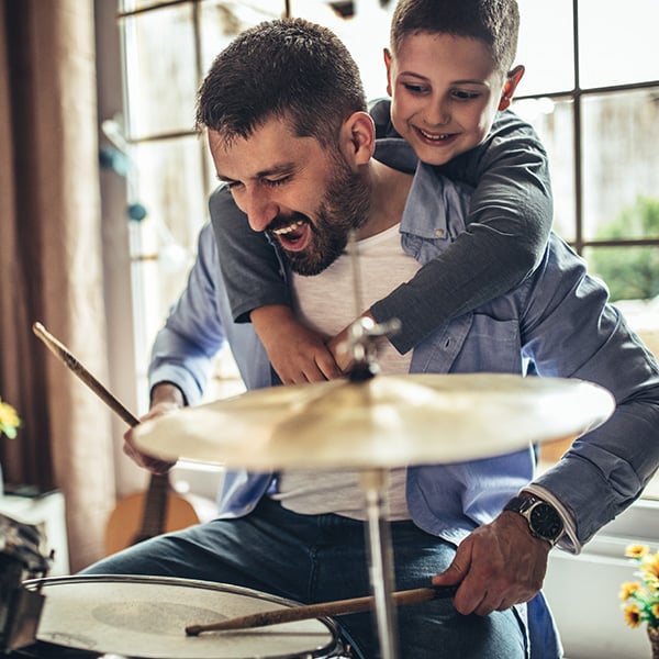 A man plays the drums while a boy hugs him from behind