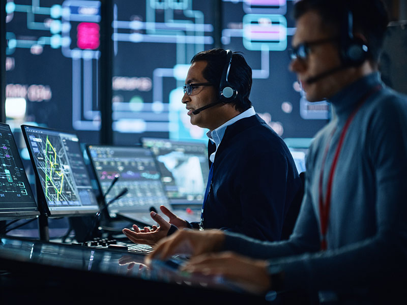 Two men work with headsets on and a large wall of screens in the background.