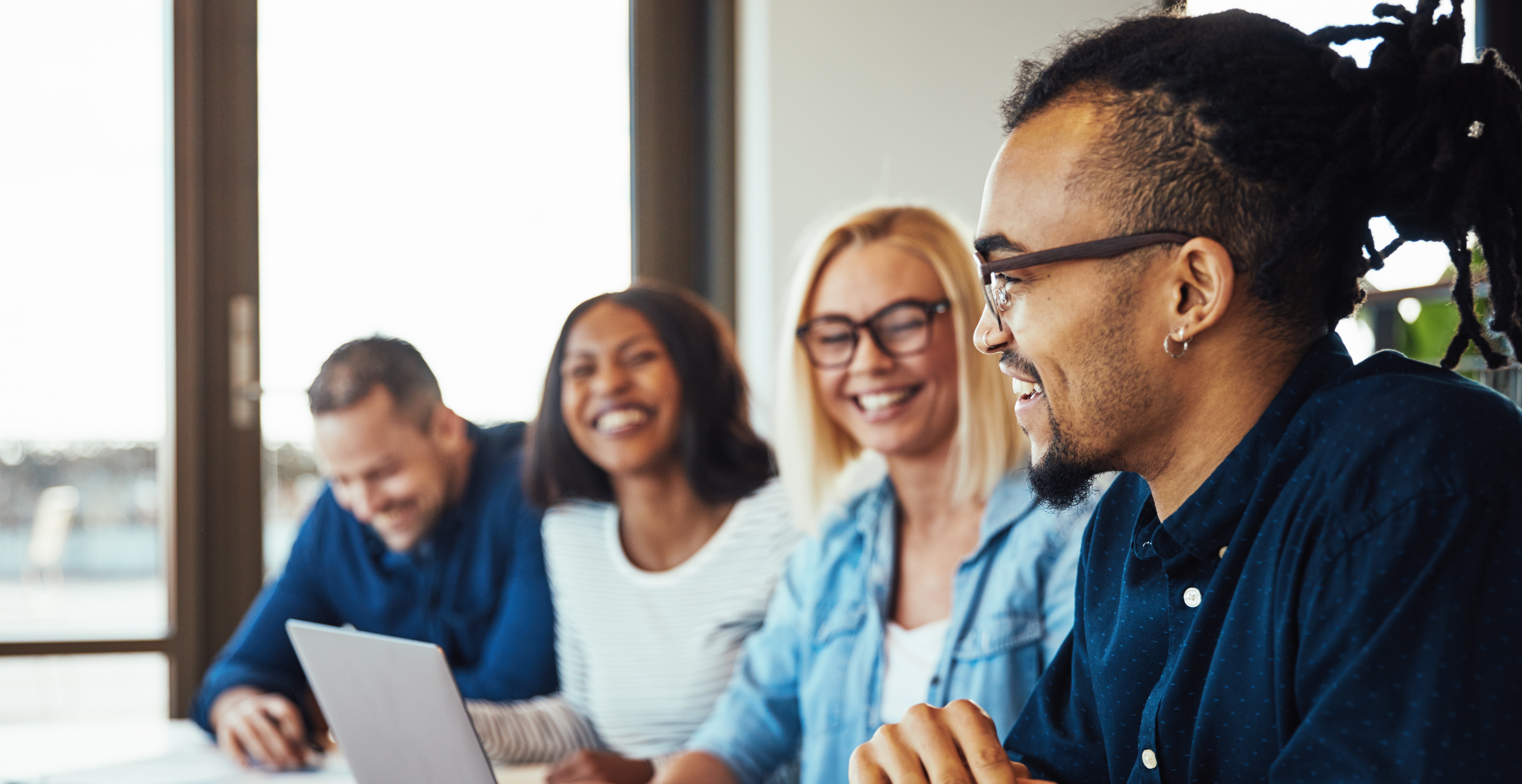 Four employees of different genders and races smile while sitting in a row at an office conference table. 