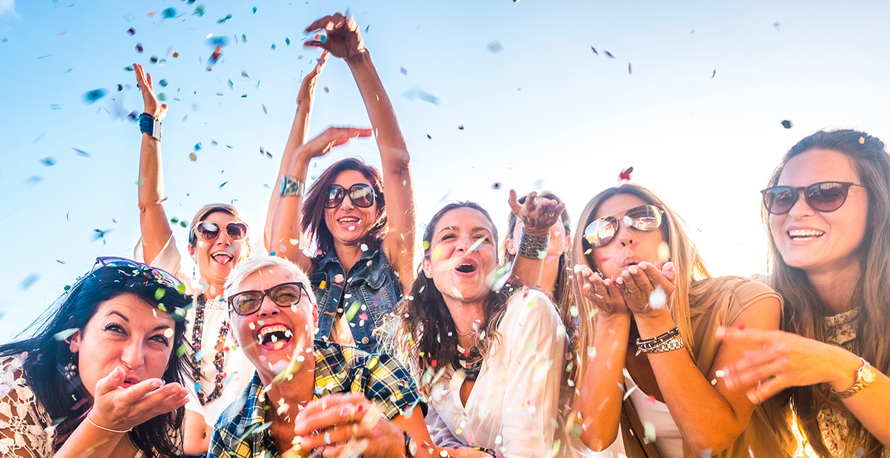 A group of women celebrate and blow confetti toward the viewer.