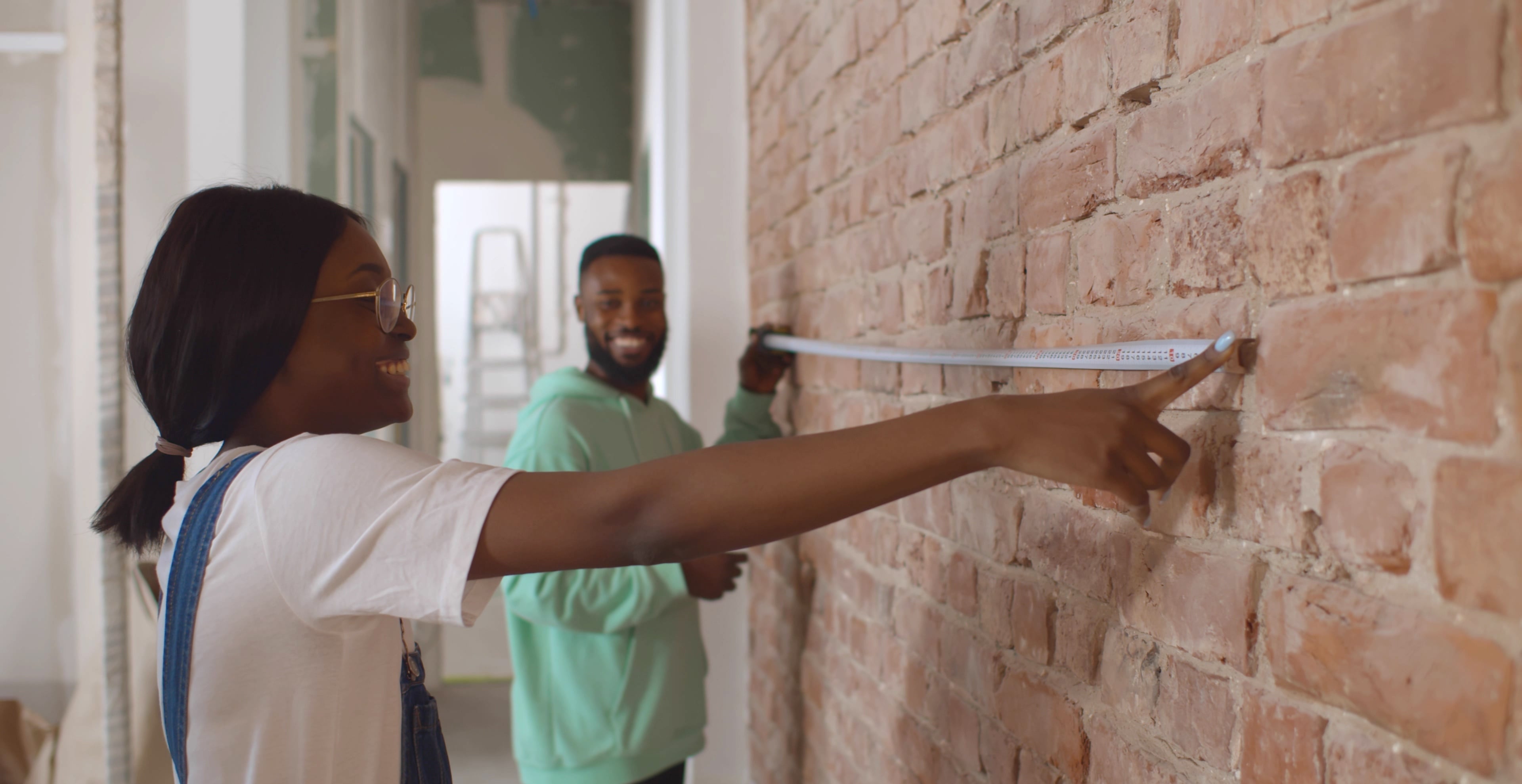 A young couple uses a tape measure against a brick wall. 