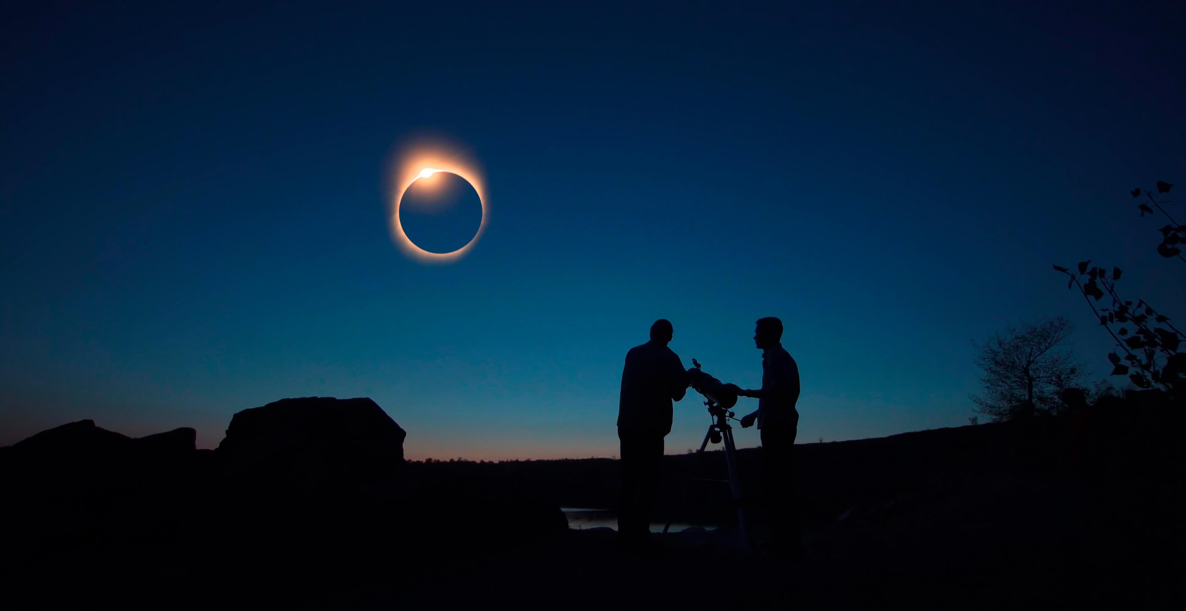 Two human silhouettes stand next to a large telescope and look toward a total solar eclipse in the sky.