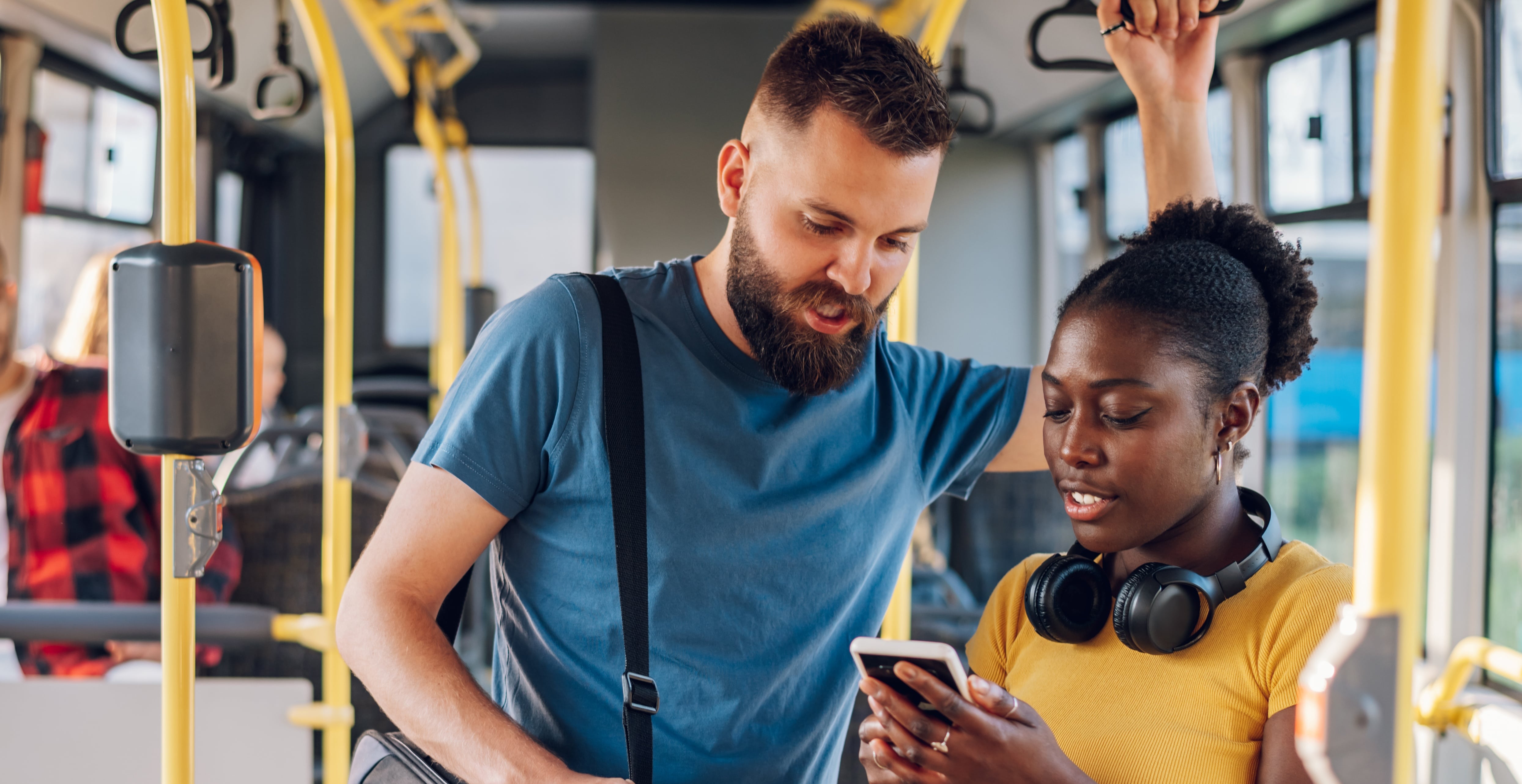 A young dark-skinned woman and a taller light-skinned man both look at a smartphone while riding a bus.  