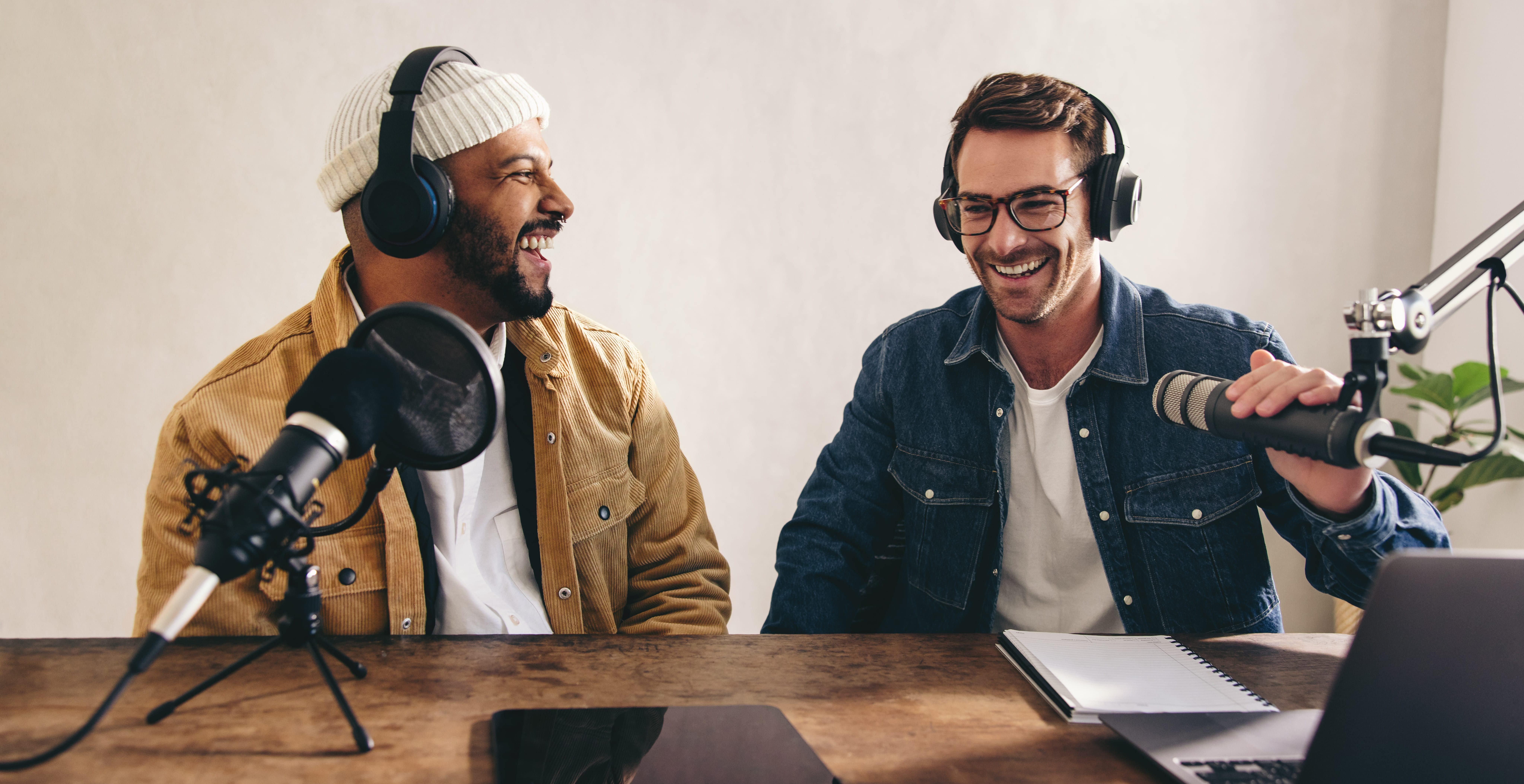 Two men wearing over-ear headphones smile as they sit at a desk set up with audio recording equipment and a laptop.