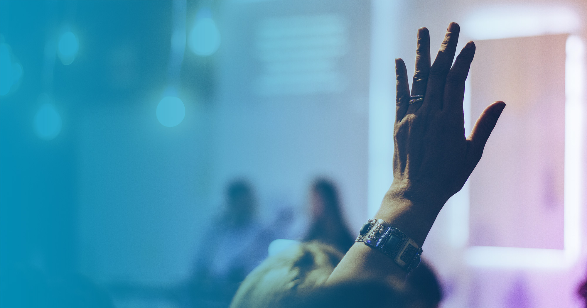 Hand raised to ask question at a conference, with speakers on stage in the background