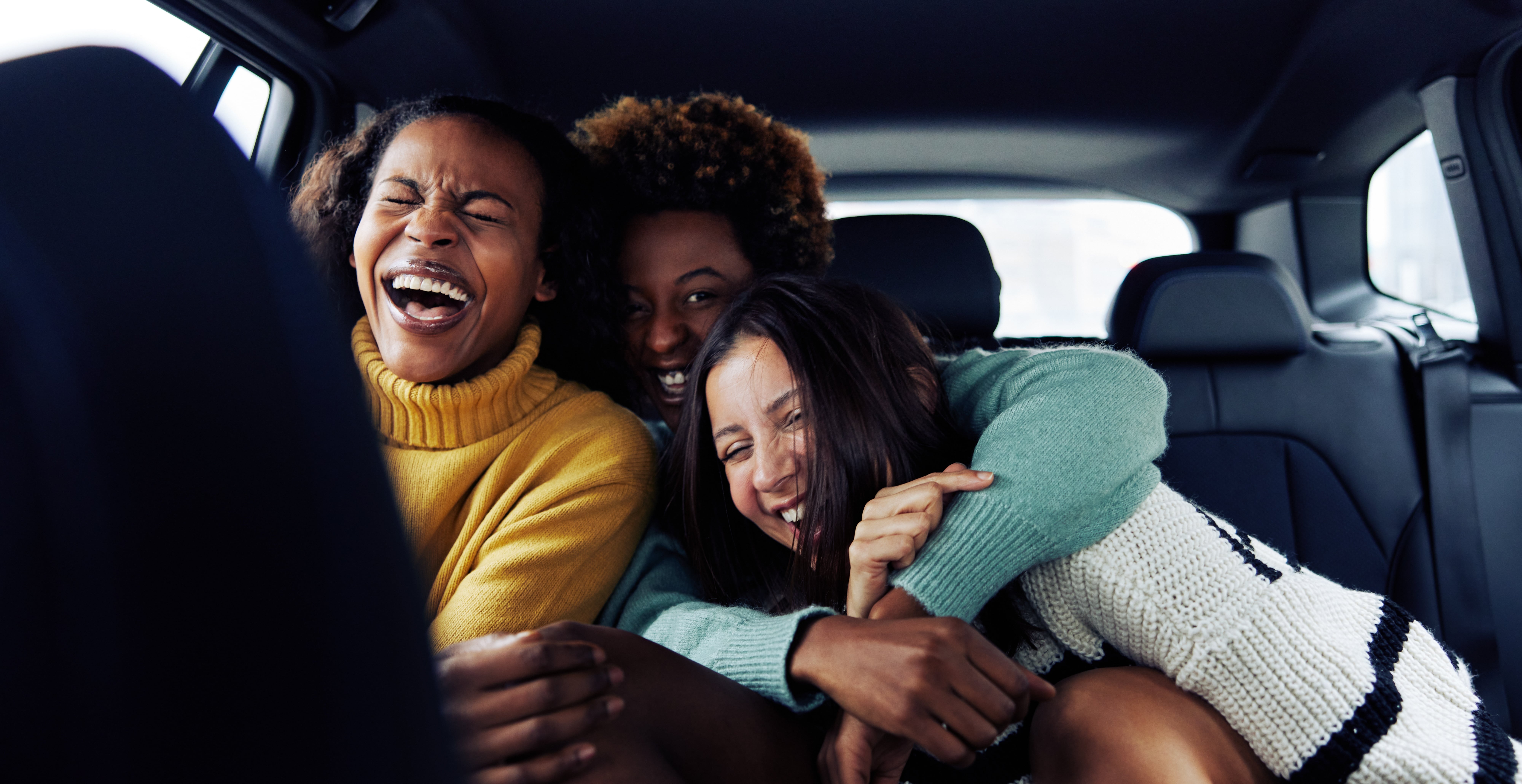 An interracial group of three women smile and laugh as they embrace in the back seat of a car. 