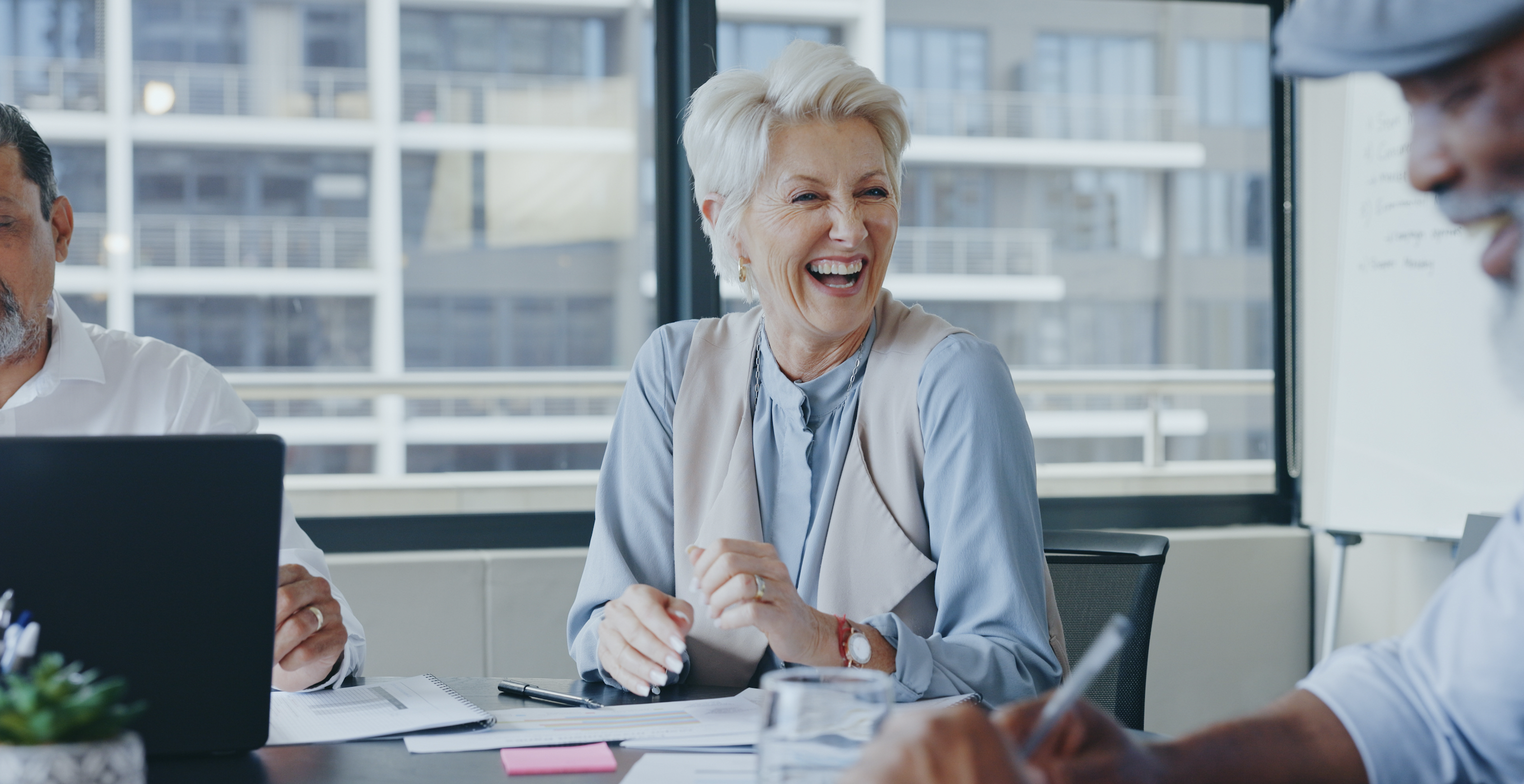 A woman with short white hair smiles while sitting at an office conference table. 