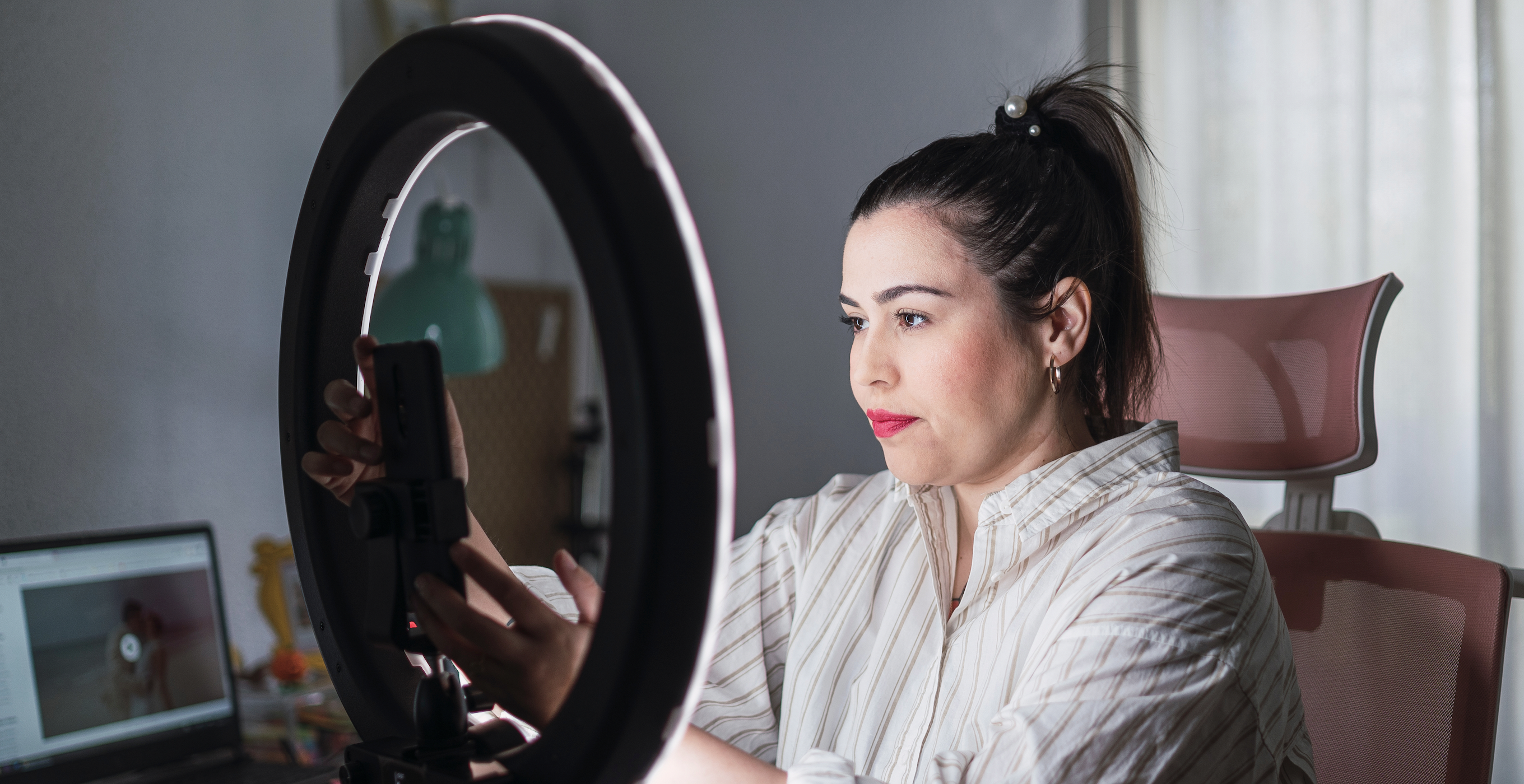 A woman with dark hair and pink lipstick adjusts a smart phone in the center of a ring light. 