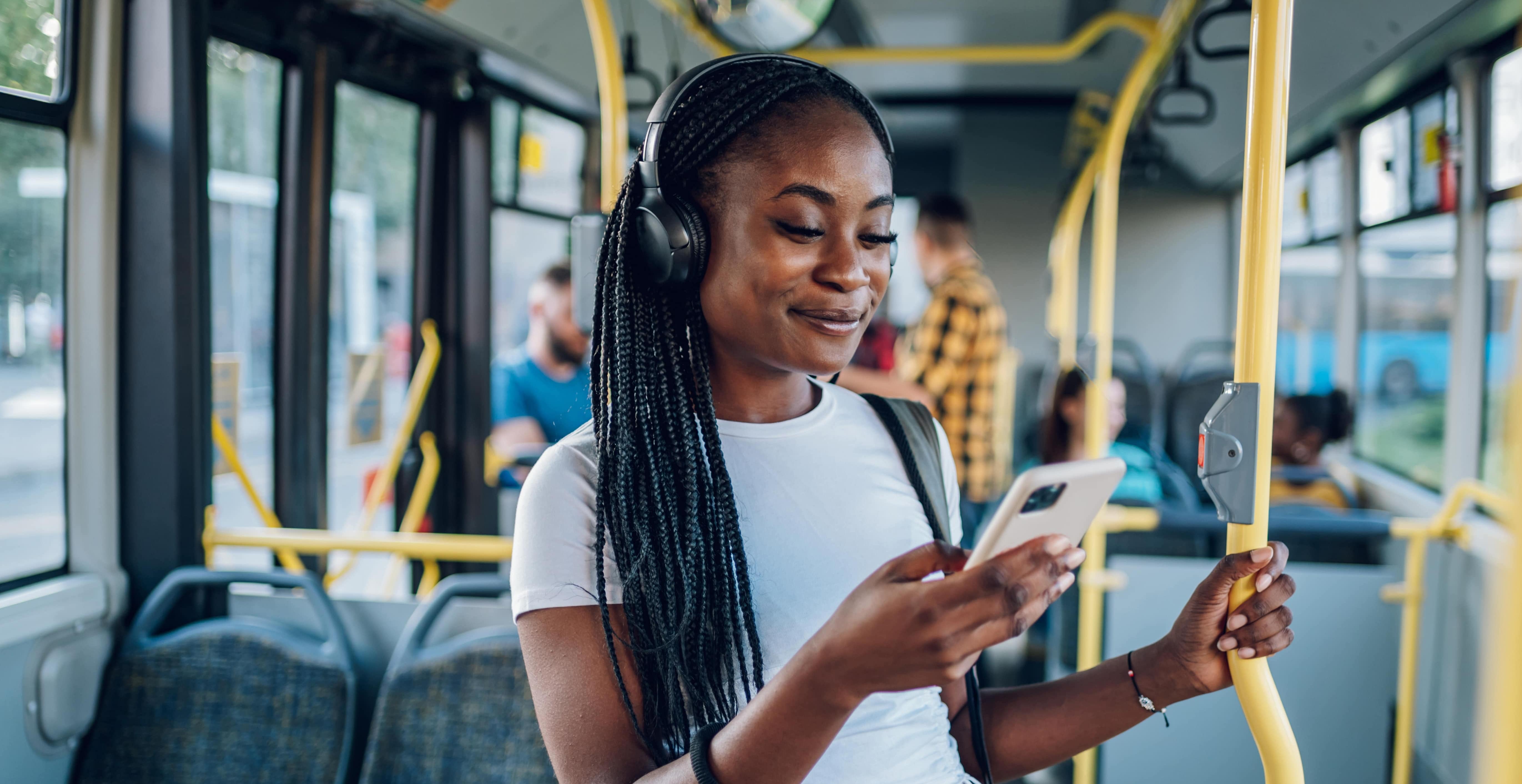 A dark-skinned young woman with long, black braids and over-the-ear headphones looks at her smartphone.