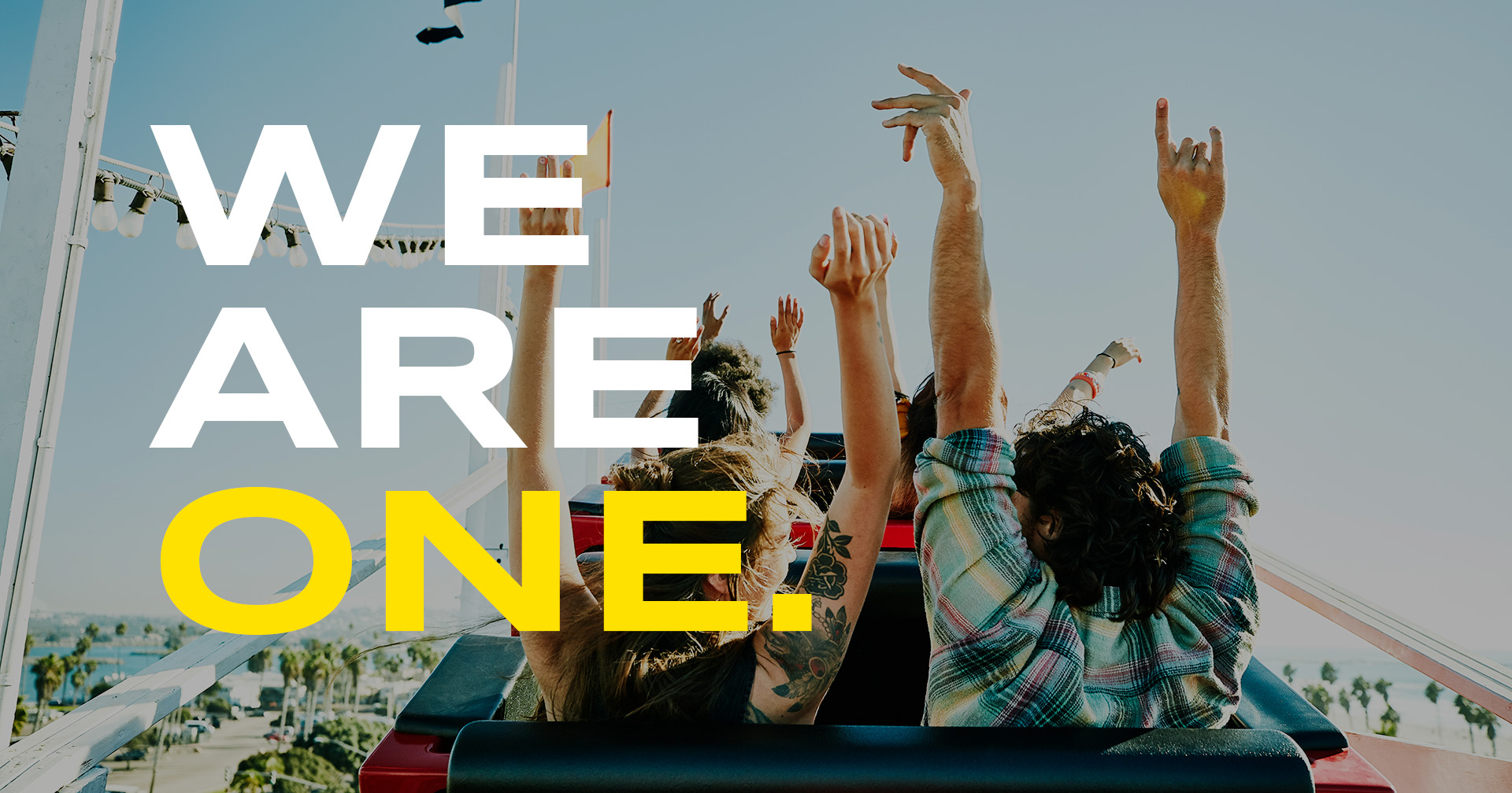 View from behind of people riding a rollercoaster and raising their hands up. The photo is overlaid with text that reads "We Are One"
