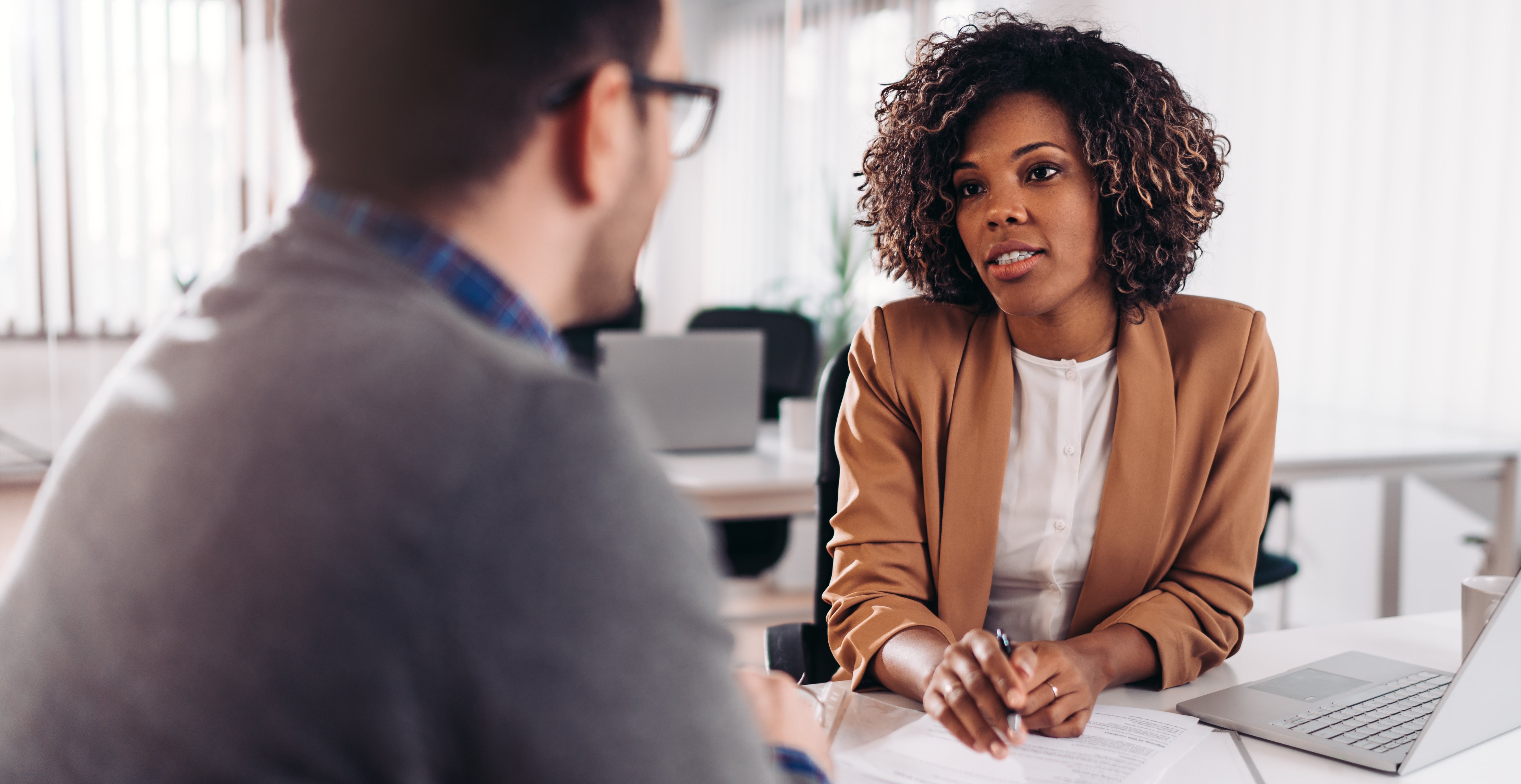 A dark-skinned woman wearing a tan blazer holds a piece of paper while speaking to a man facing toward her. 