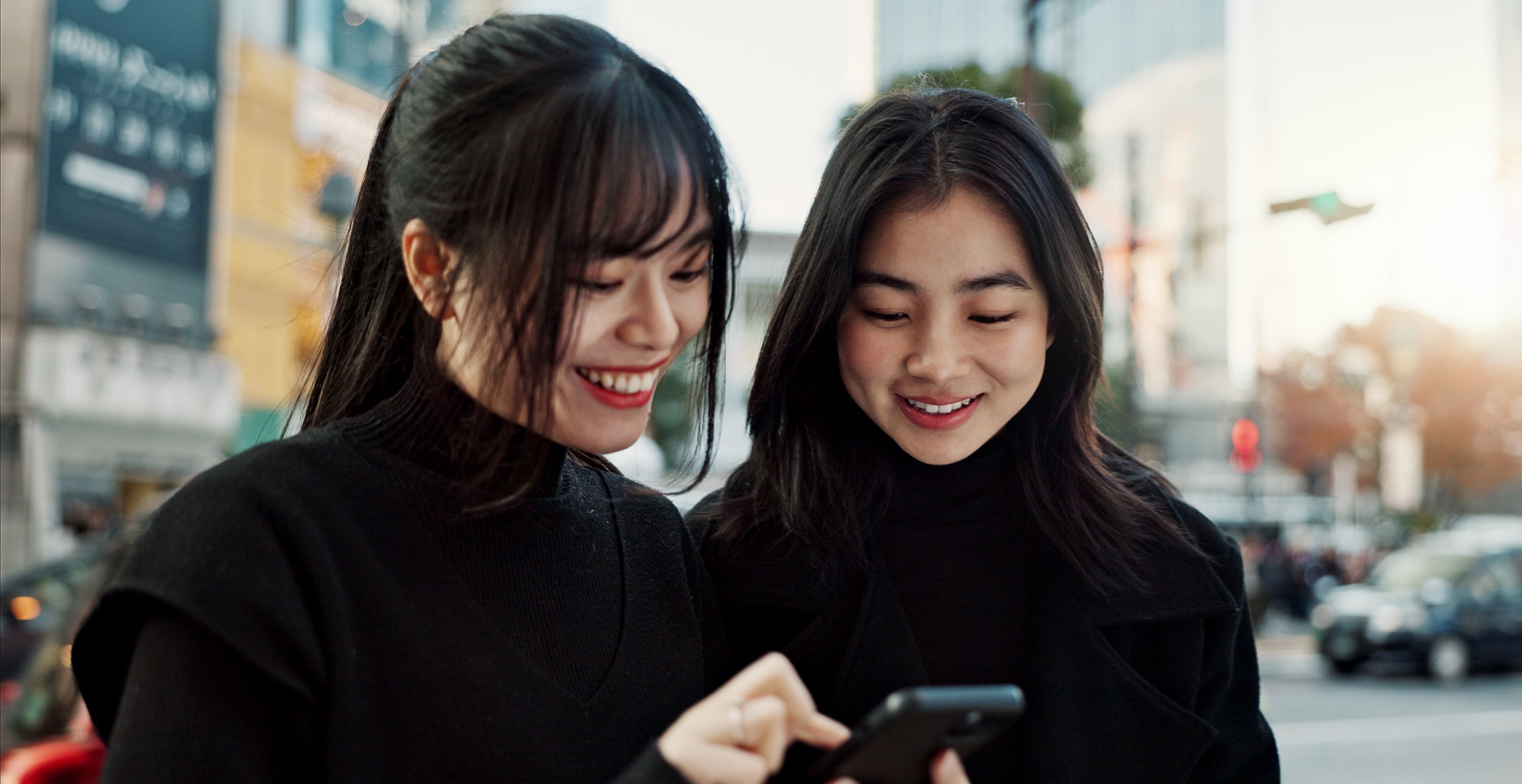 Two young women smile while looking at a smartphone screen together. 