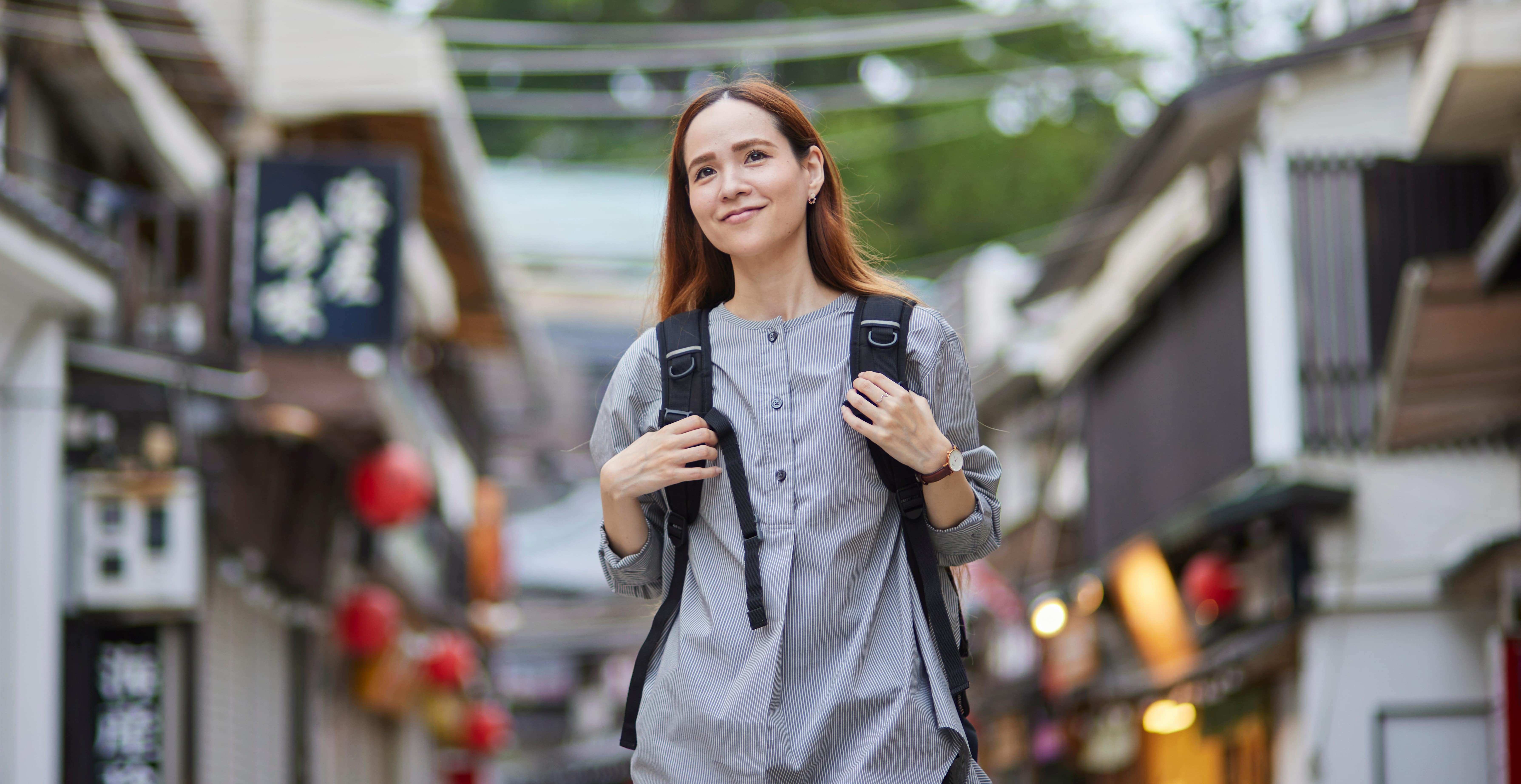 A light-skinned young woman with long, brown hair carries a backpack while walking on the street.