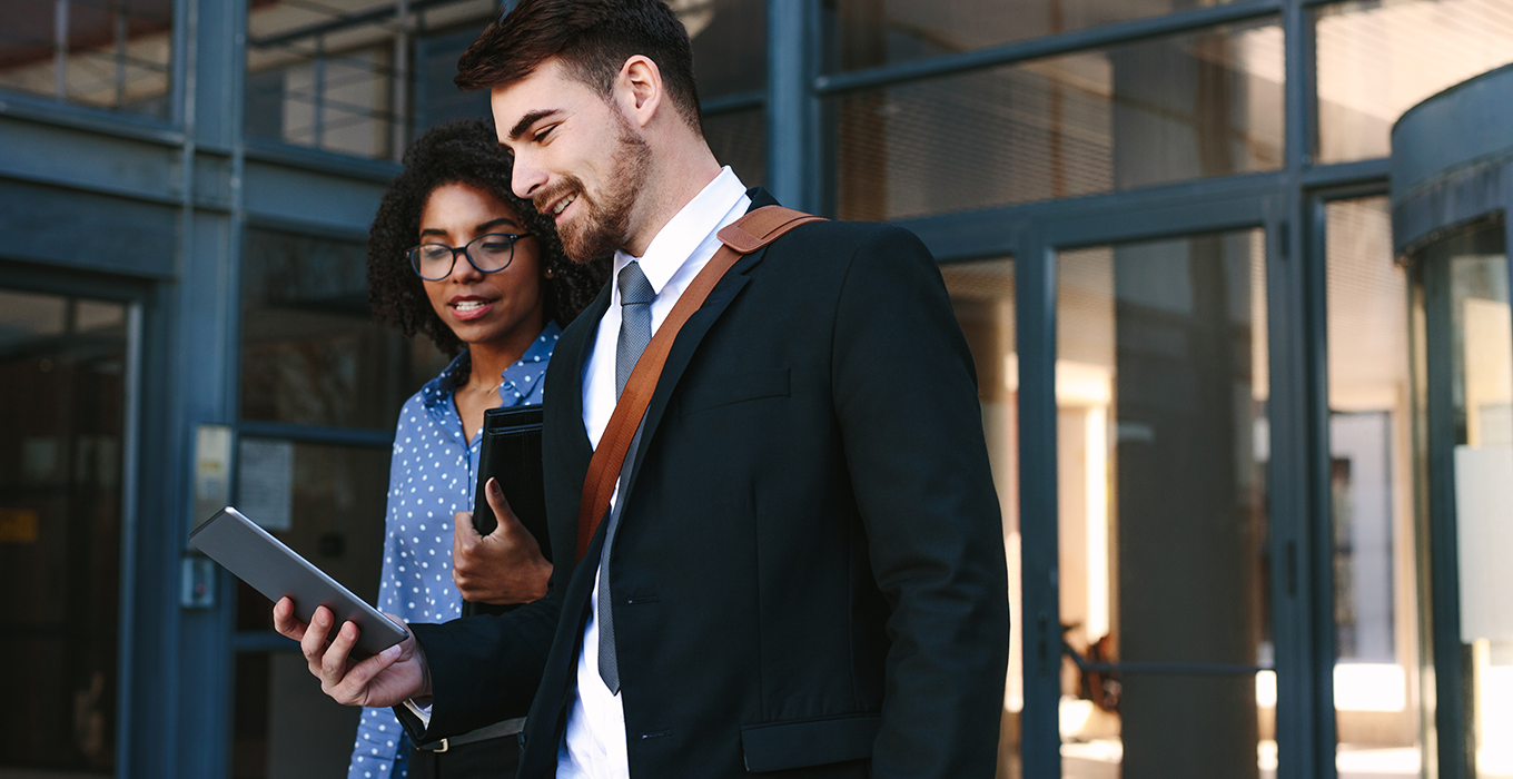 Two young professionals exit a building while both looking at a tablet device.